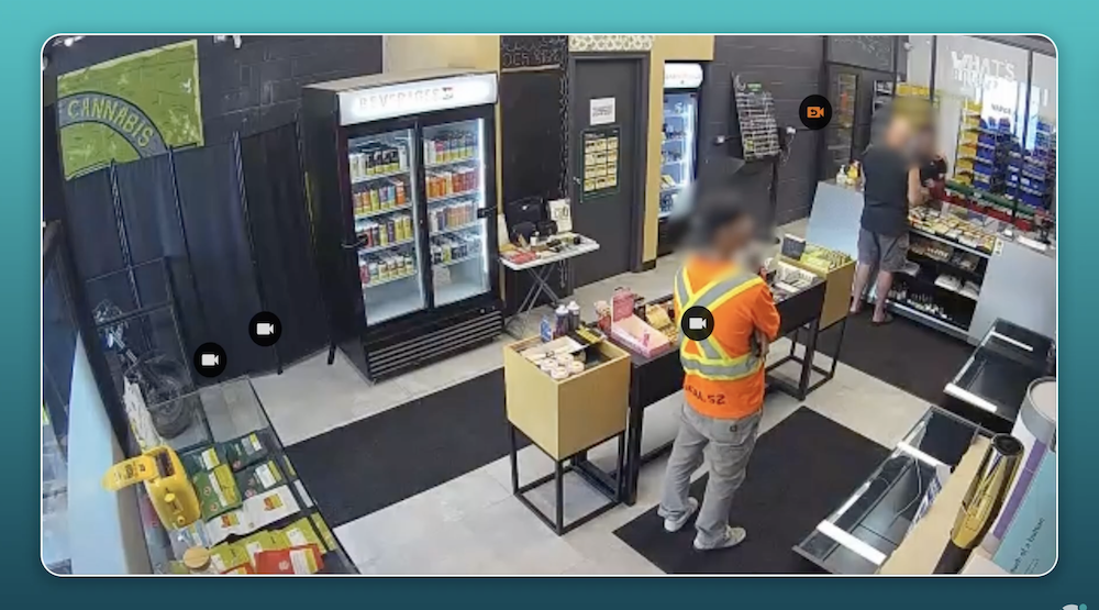 Two people are inside a cannabis store; one browses near shelves while the other, wearing a high-visibility vest, stands by a central display.