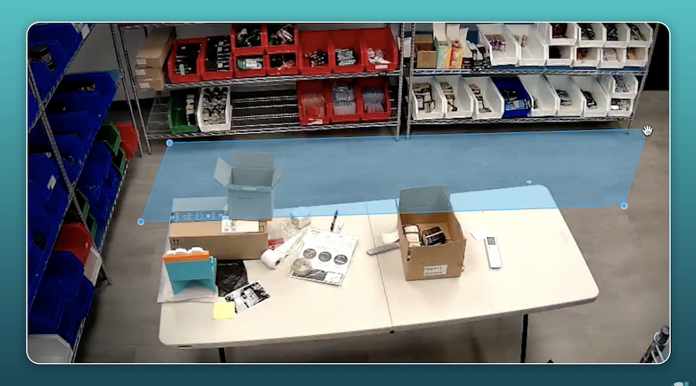 A worktable with open boxes, tools, and various items sits in front of shelves filled with organized bins in a storage or packaging area.