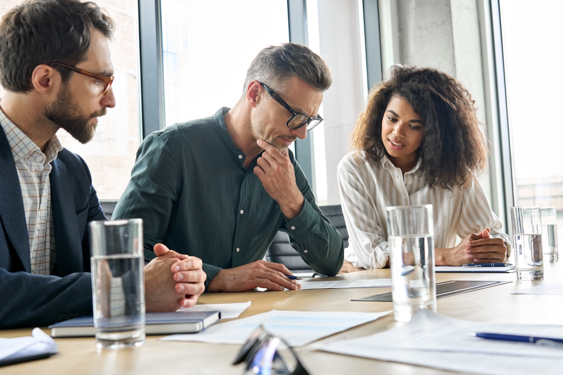 Three people sit at a conference table with papers, notebooks, and glasses of water, engaged in a business meeting or discussion.