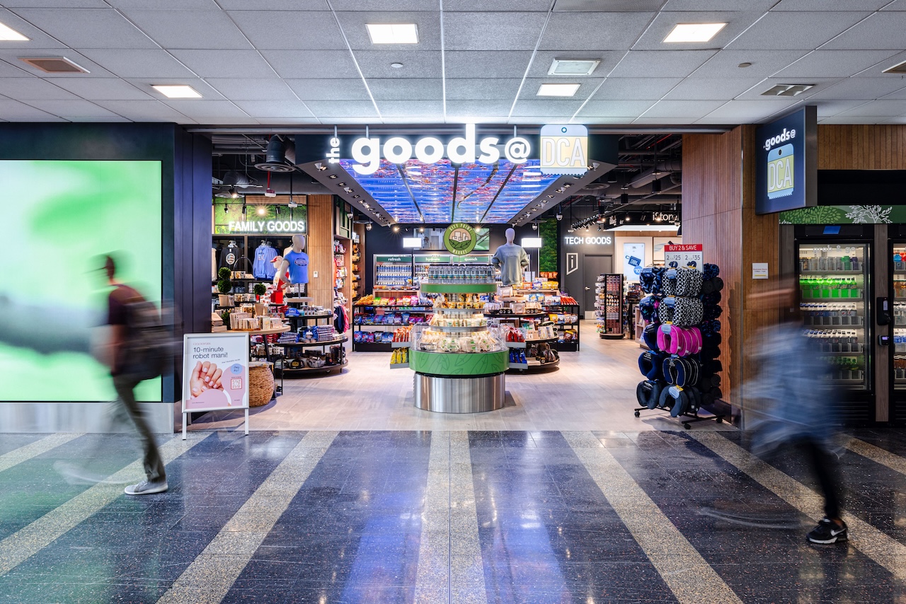 Travelers walk past a brightly lit airport convenience store named "goods@DCA," displaying snacks, drinks, and travel essentials near its entrance.