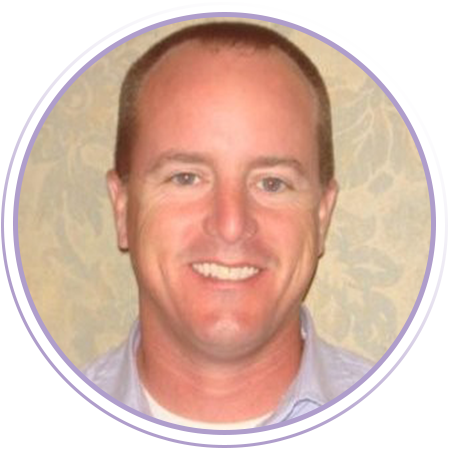 A man with short brown hair smiles at the camera, wearing a light purple collared shirt, with a patterned background.