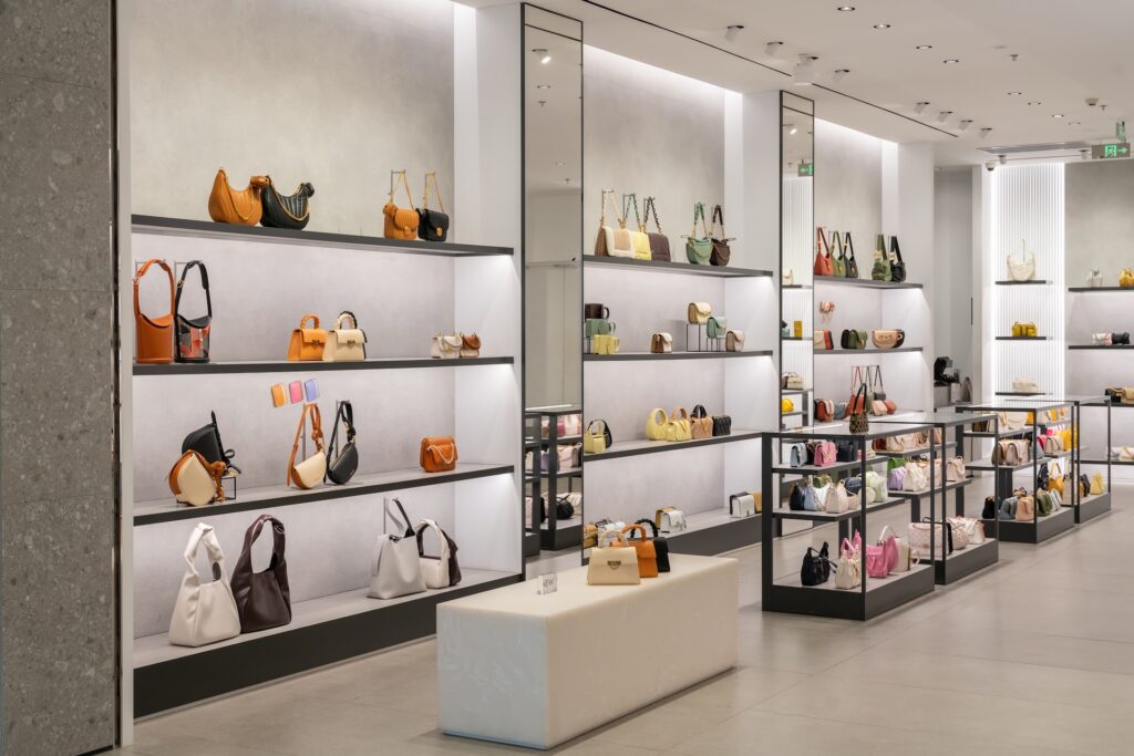 Modern handbag store interior with illuminated shelves and glass displays showcasing various purses and handbags in different colors and styles, with a bench in the center.