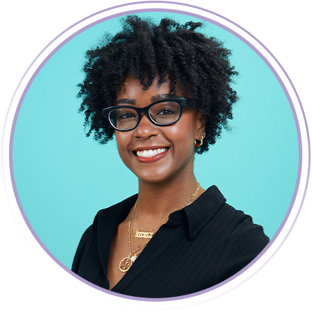 A smiling woman with curly hair and glasses, wearing a black top and layered necklaces, poses in front of a light blue background.