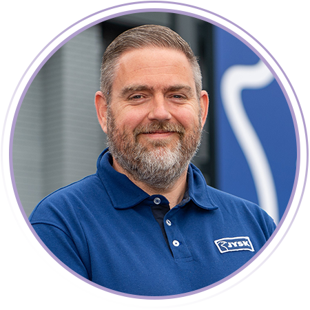 A man with short brown hair and a beard, wearing a blue collared shirt with a "KWB" logo, stands in front of a blurred blue and gray background.