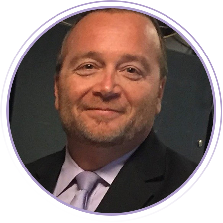 A middle-aged man with short hair and a beard, wearing a suit and light purple tie, is smiling at the camera.