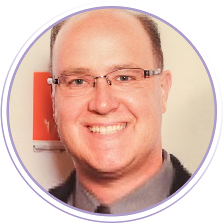 A man with short hair and glasses smiles at the camera, wearing a gray shirt and dark jacket, with a blurred red and white background.