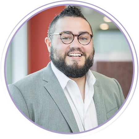 A person wearing glasses and a gray blazer over a white shirt smiles at the camera in an indoor setting with blurred background.