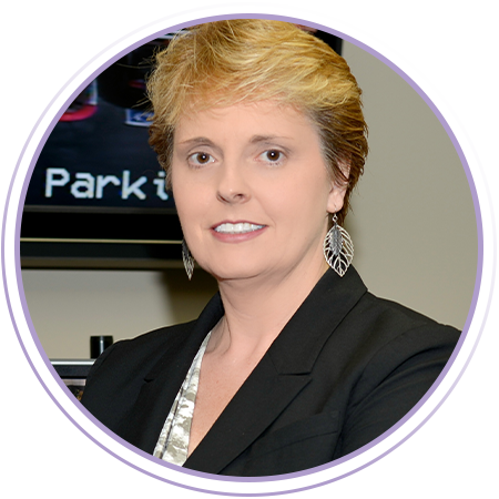 A woman with short blond hair, wearing a black blazer, silver leaf earrings, and a light patterned blouse, stands in front of a blurred background with partial text visible.