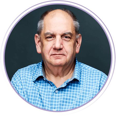 A middle-aged man with short gray hair, wearing a blue checkered shirt, looks directly at the camera against a dark background.