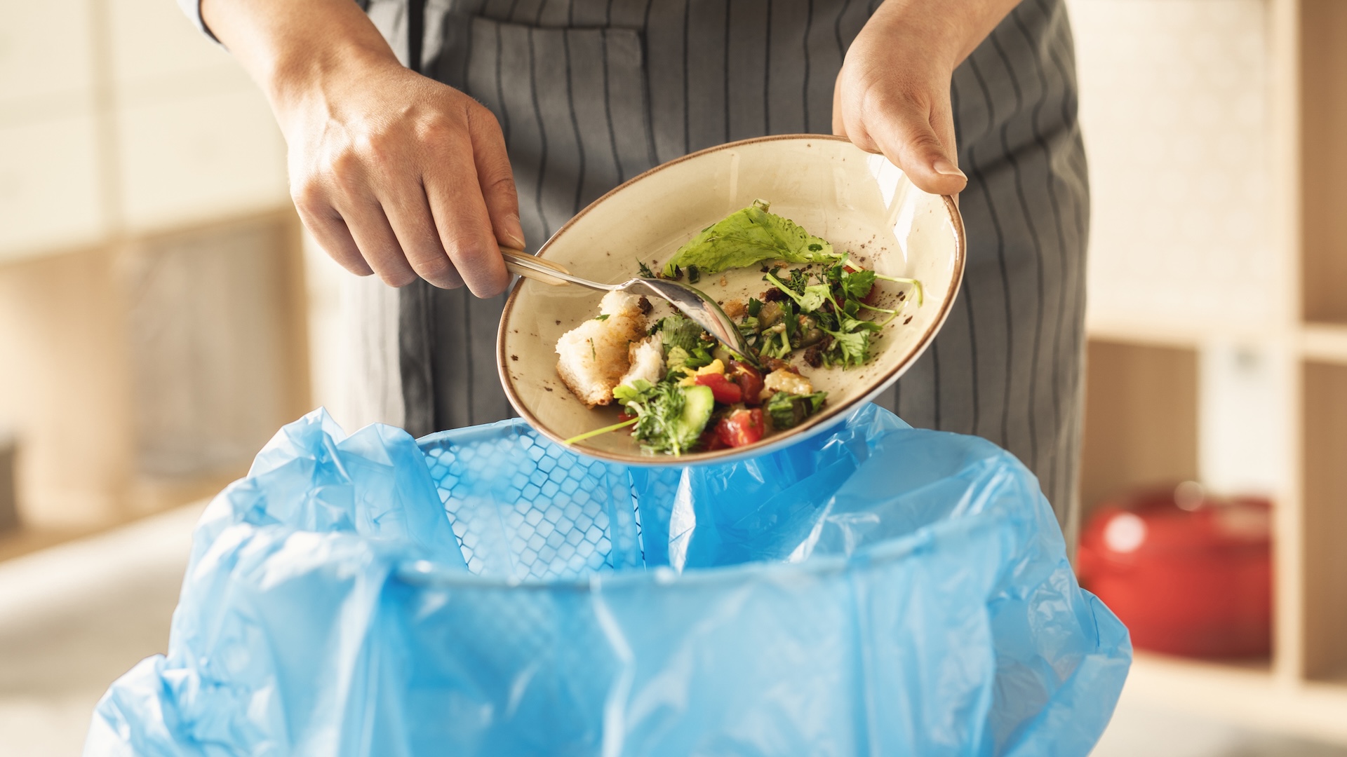 A person scrapes leftover food from a plate into a trash bin lined with a blue plastic bag.