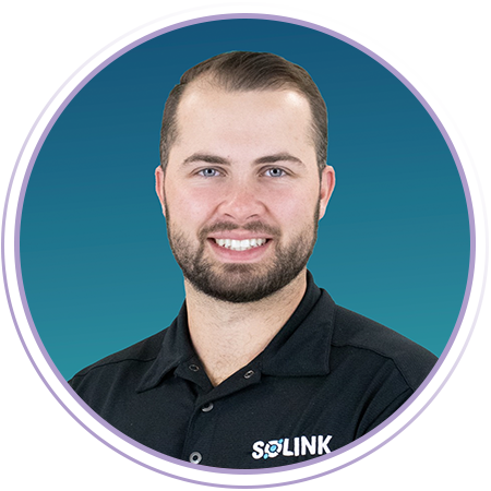 A man with short brown hair and a beard, wearing a black SOLINK polo shirt, smiling in front of a blue gradient background.