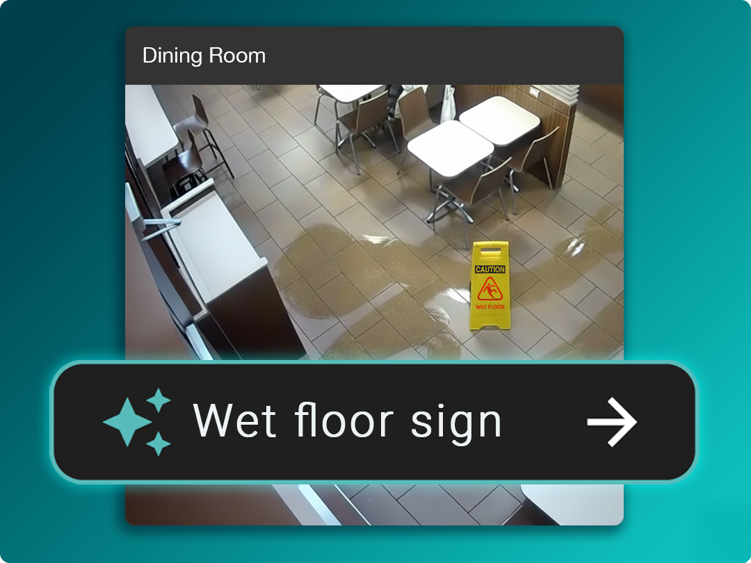 A wet floor caution sign stands on a tiled floor in a dining area, with visible water puddles nearby and tables and chairs in the background.