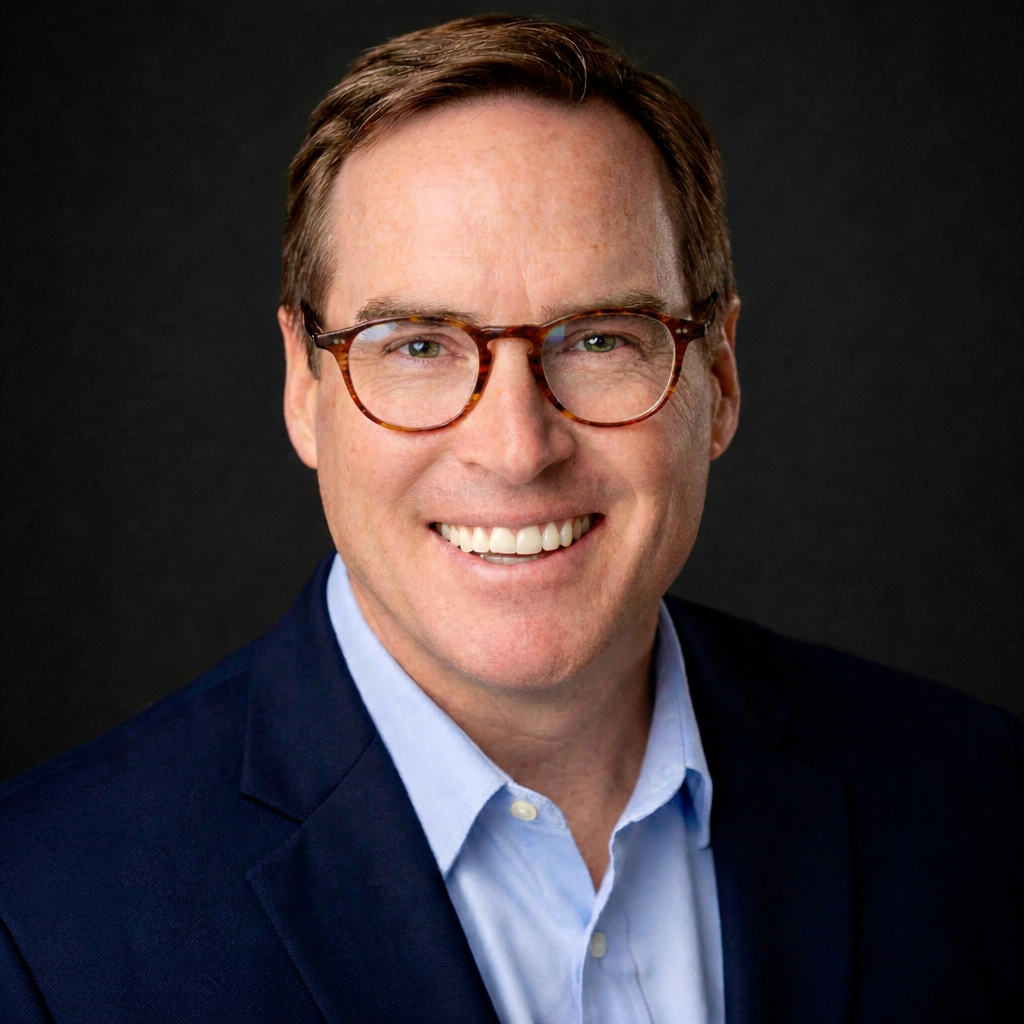 A man with short brown hair, wearing glasses, a navy blazer, and a light blue shirt, smiles at the camera against a dark background.