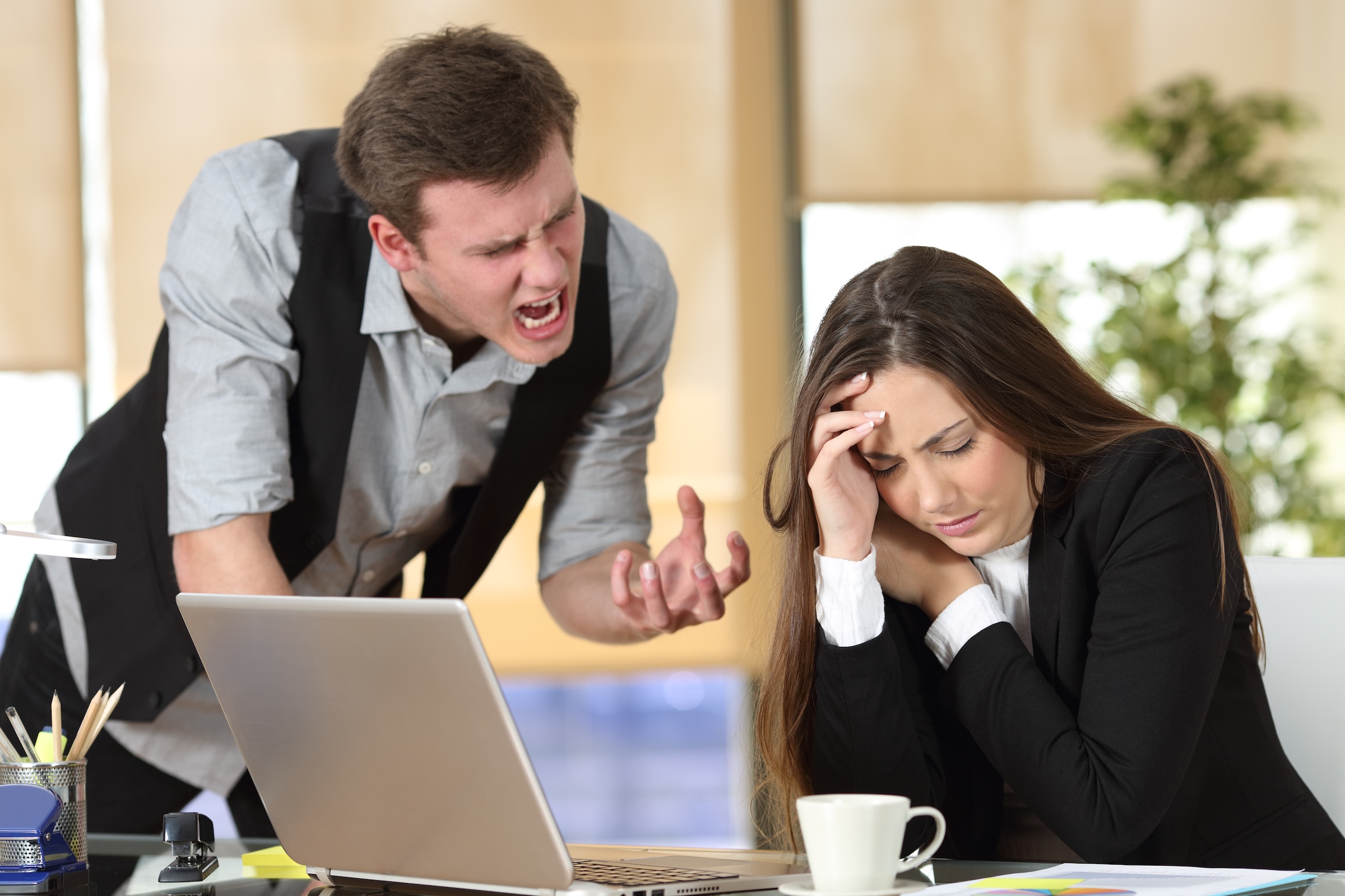 A man standing and angrily yelling at a woman who looks stressed and holds her head in her hand while sitting at a desk with a laptop and papers.