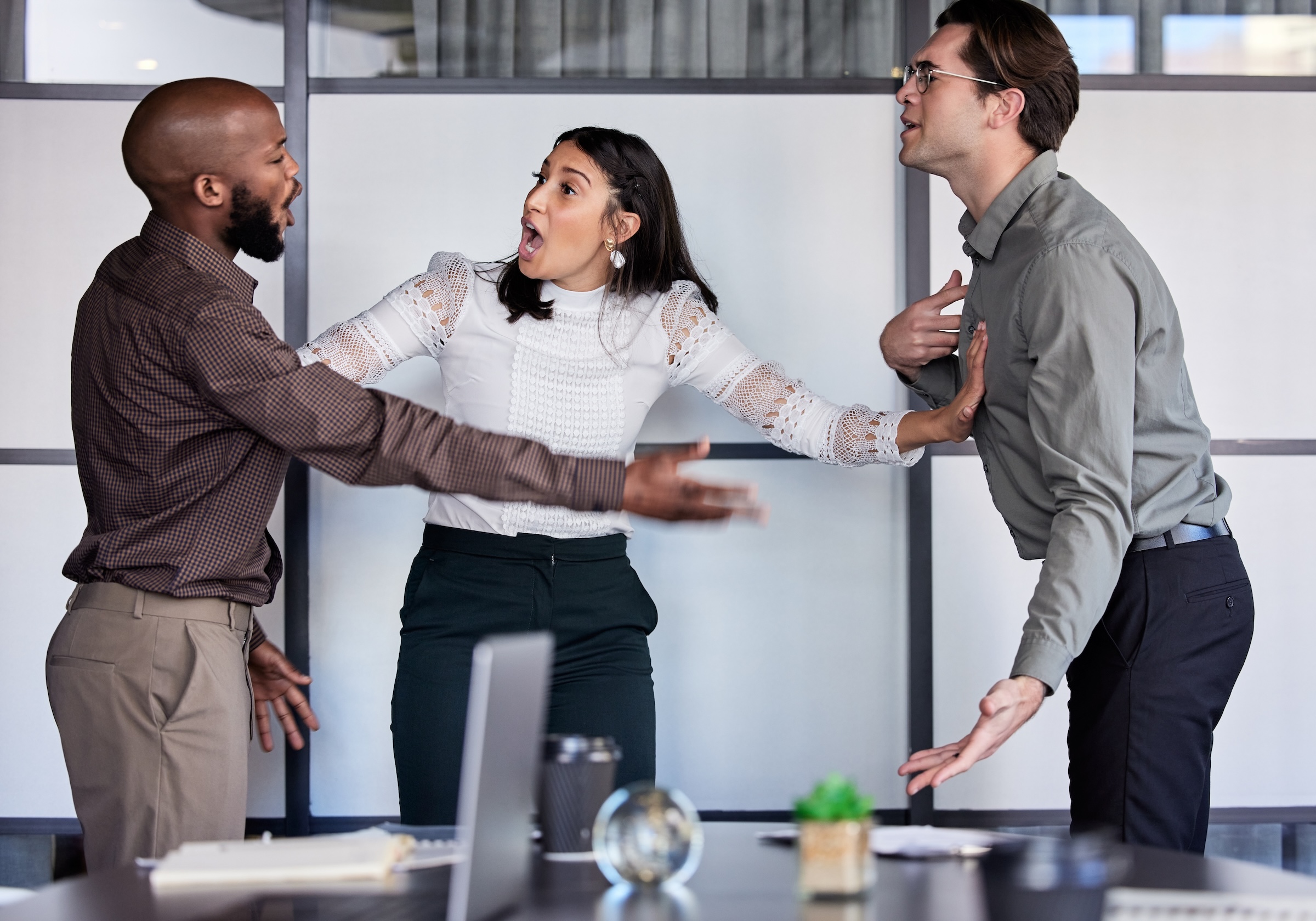 Three coworkers in an office appear to argue, with one woman standing between two men, raising her arms to separate them.