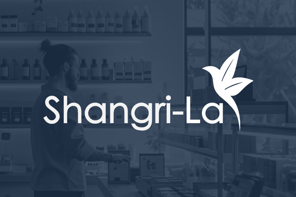 Man with a beard stands at a counter in a modern store; overlaid text reads “Shangri-La” with a stylized bird logo beside it.